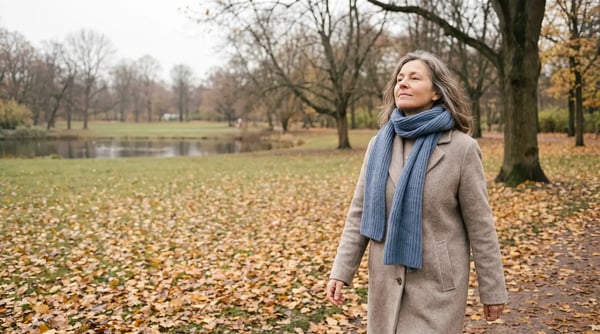 Frau um die 50 mit Wollmantel und blauem Schal geht an einem Herbsttag durch einen Park mit buntem Laub